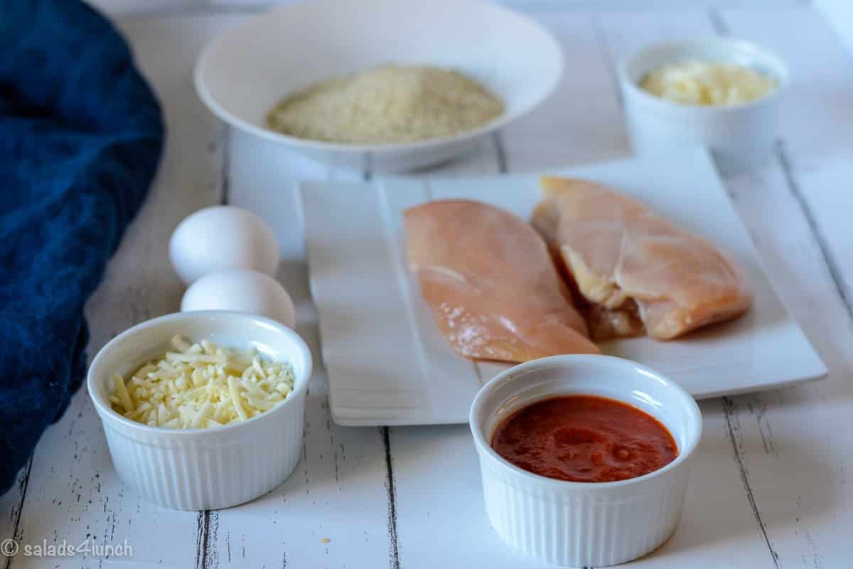 Photo showing ingredients needed to make Air Fryer Chicken Parmesan: Italian bread crumbs, parmesan cheese, chicken breasts, two eggs, mozzarella cheese and marinara sauce.