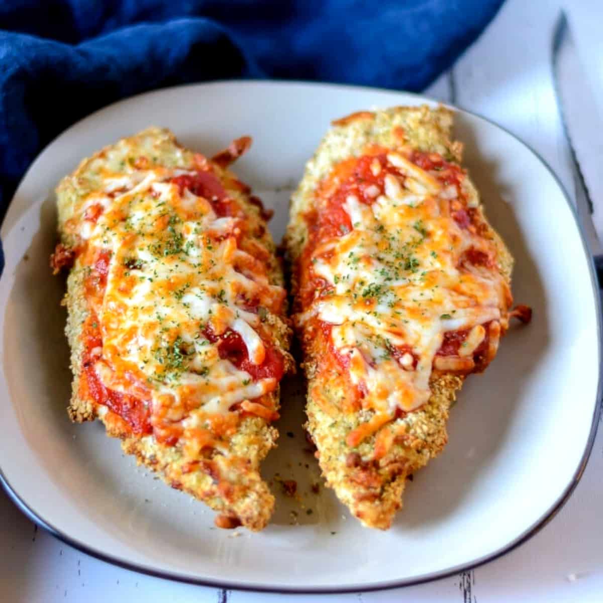 Square photo of two chicken parmesan on a square white plate with a blue napkin in the background.
