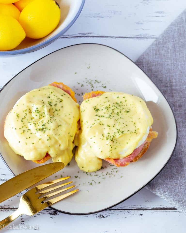 Close up photo of two delicious eggs benedict on a white plate with black trim on a grey napkin with a gold fork and knife.