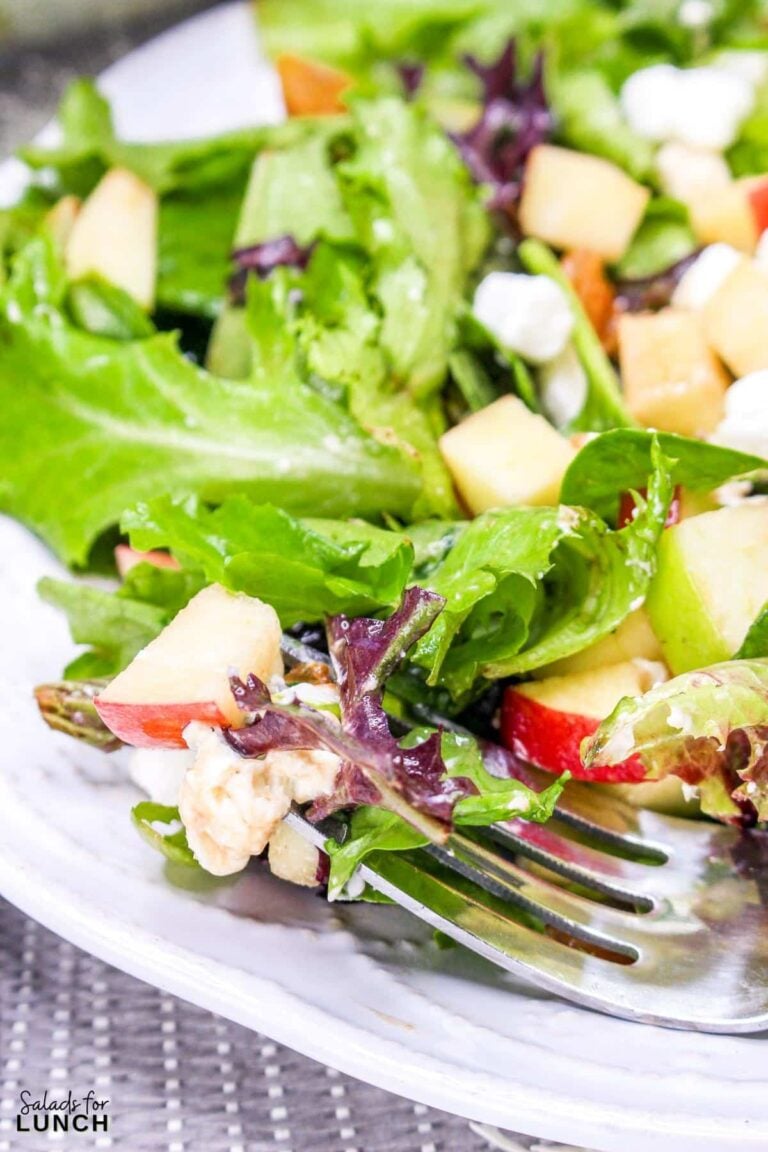 Close up of a mixed greens salad with apple, goat cheese, raisins and toasted walnuts on a white plate with a fork.