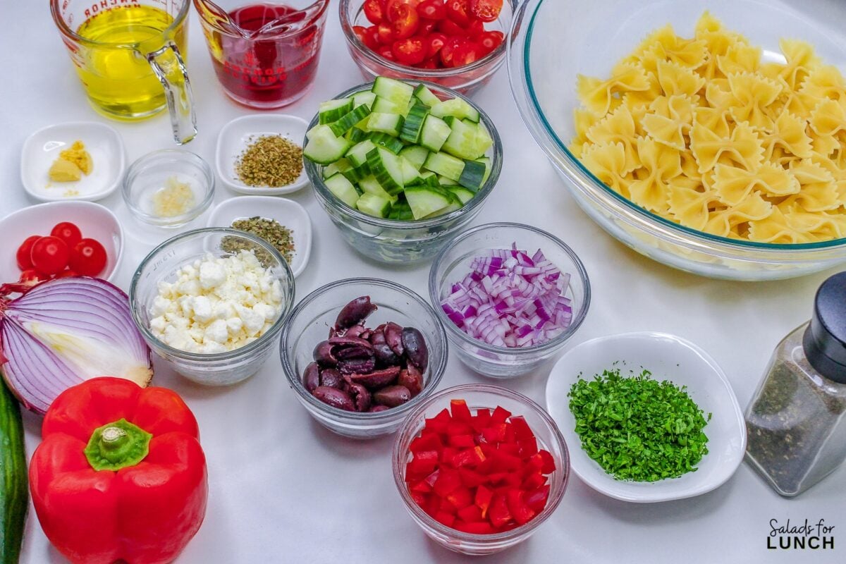 Ingredients for greek pasta salad in small bowls including: red pepper, black olives, cucumber, red onion, bow tie pasta, olive oil, red wine vinegar, herbs and spices.