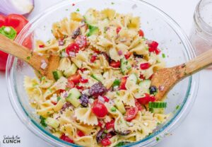 Glass bowl with cucumber, kalamata olives, tomatoes, red onion, feta cheese, parsley, and red bell pepper mixed together with bow tie pasta.