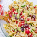 Close up of a glass bowl with cucumber, kalamata olives, tomatoes, red onion, feta cheese, parsley, and red bell pepper mixed together with bow tie pasta.