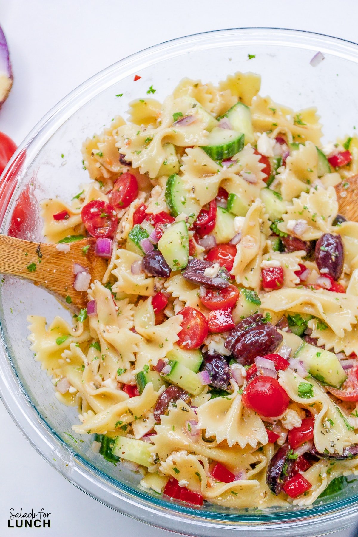 Close up of a glass bowl with cucumber, kalamata olives, tomatoes, red onion, feta cheese, parsley, and red bell pepper mixed together with bow tie pasta.