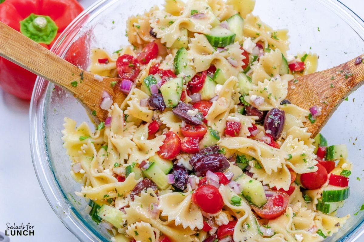 Close up of a glass bowl with cucumber, kalamata olives, tomatoes, red onion, feta cheese, parsley, and red bell pepper mixed together with bow tie pasta.