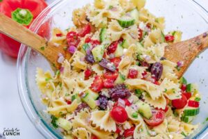 Close up of a glass bowl with cucumber, kalamata olives, tomatoes, red onion, feta cheese, parsley, and red bell pepper mixed together with bow tie pasta.