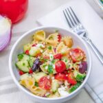 Close up of a white bowl full of bow tie pasta with cucumbers, tomato, olives, feta cheese and greek salad dressing.