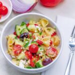 Close up of a white bowl full of bow tie pasta with cucumbers, tomato, olives, feta cheese and greek salad dressing.