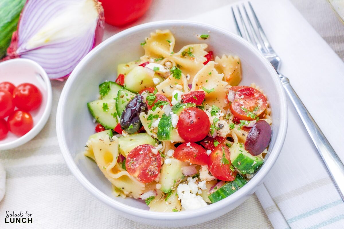 Close up of a white bowl full of bow tie pasta with cucumbers, tomato, olives, feta cheese and greek salad dressing.