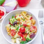 Close up of a white bowl full of bow tie pasta with cucumbers, tomato, olives, feta cheese and greek salad dressing.