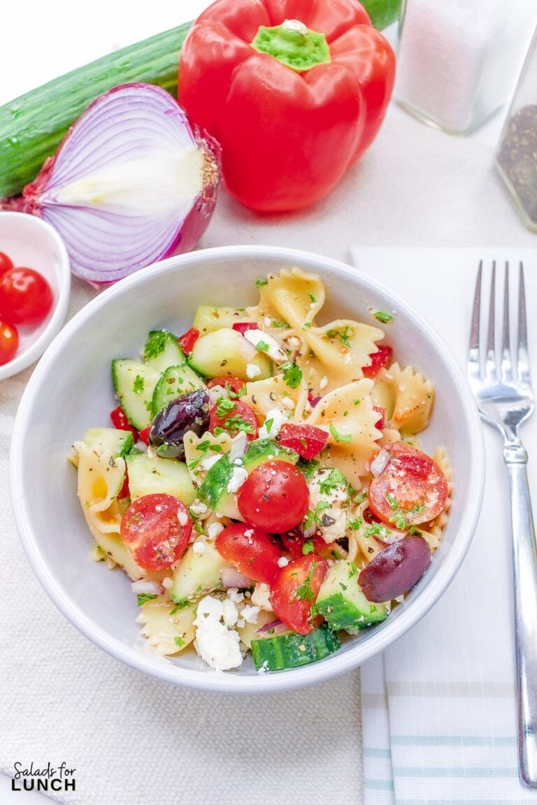Close up of a white bowl full of bow tie pasta with cucumbers, tomato, olives, feta cheese and greek salad dressing.