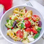 Close up of a white bowl full of bow tie pasta with cucumbers, tomato, olives, feta cheese and greek salad dressing.