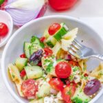 Close up of a white bowl full of bow tie pasta with cucumbers, tomato, olives, feta cheese and greek salad dressing.