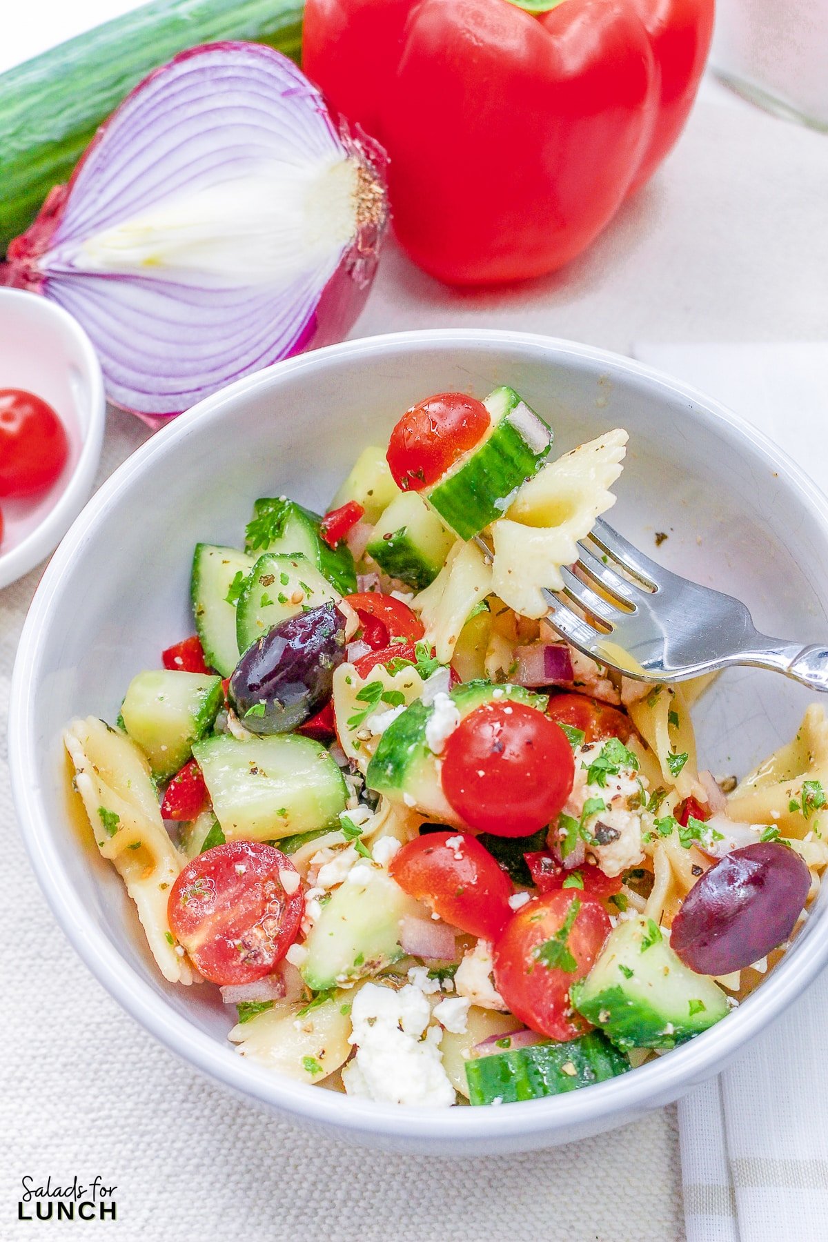Close up of a white bowl full of bow tie pasta with cucumbers, tomato, olives, feta cheese and greek salad dressing.