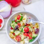 White bowl full of bow tie pasta with cucumbers, tomato, olives, feta cheese and greek salad dressing.