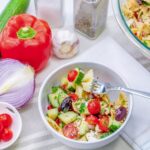White bowl full of bow tie pasta with cucumbers, tomato, olives, feta cheese and greek salad dressing.