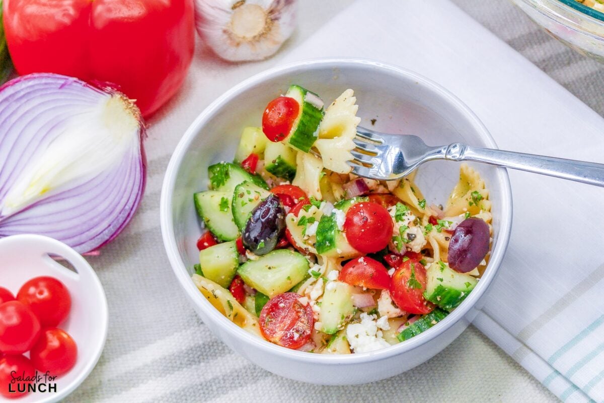 White bowl full of bow tie pasta with cucumbers, tomato, olives, feta cheese and greek salad dressing.