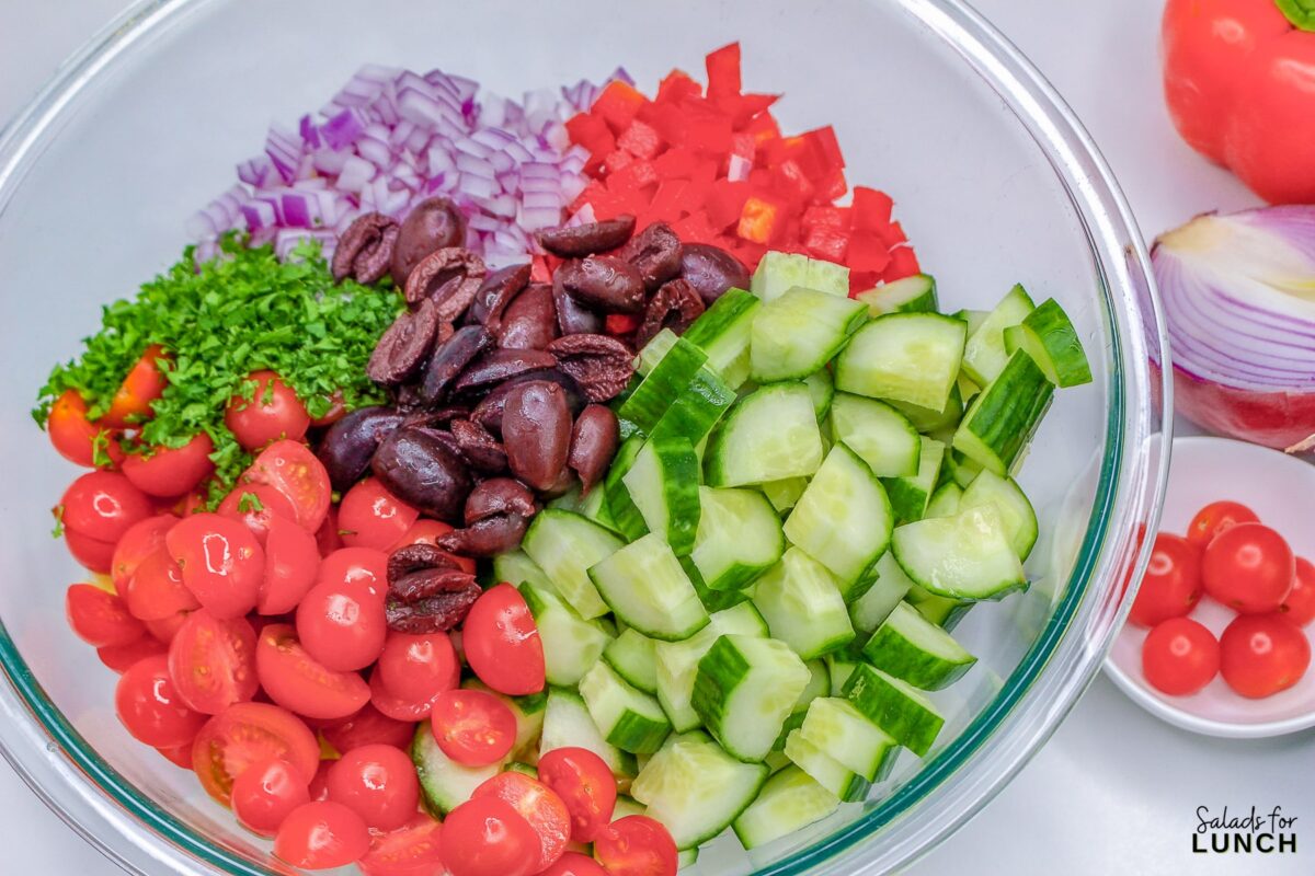 Glass bowl with cucumber, kalamata olives, tomatoes, red onion, feta cheese, parsley, and red bell pepper.