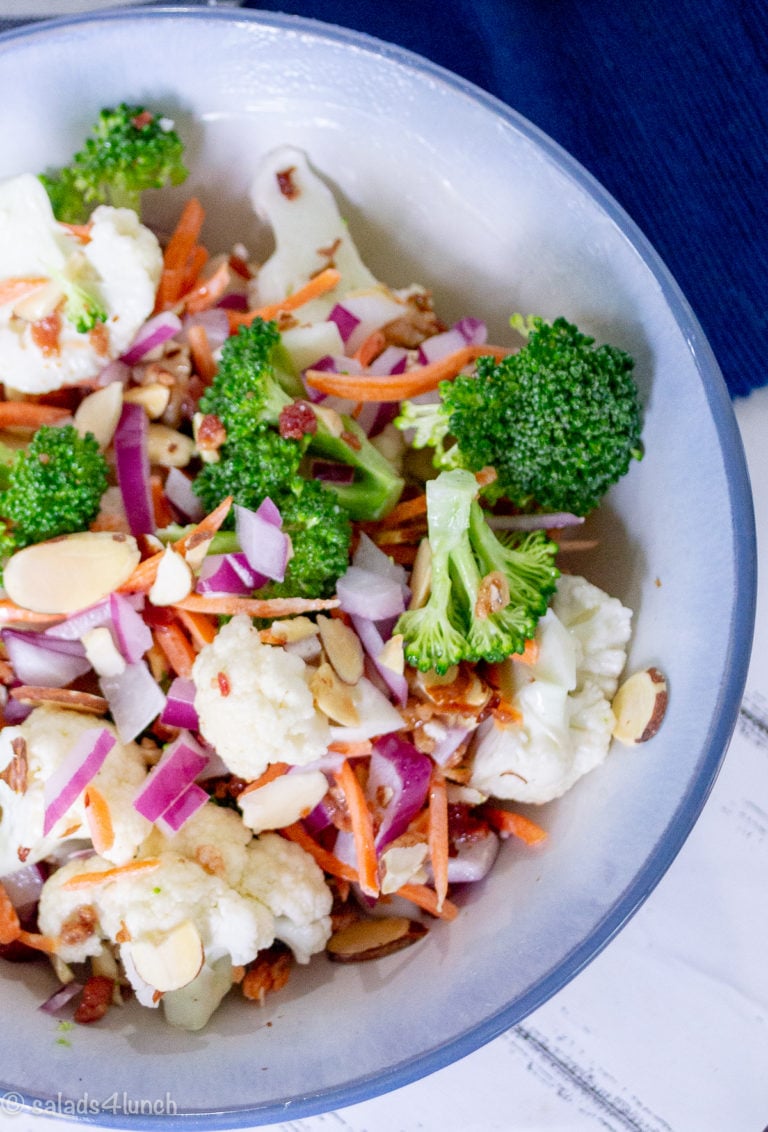 Close up of broccoli cauliflower salad with almonds, carrots and bacon in a white bowl with blue trim on a blue placemat with text overlay"Broccoli Cauliflower Salad with Bacon and Almonds"