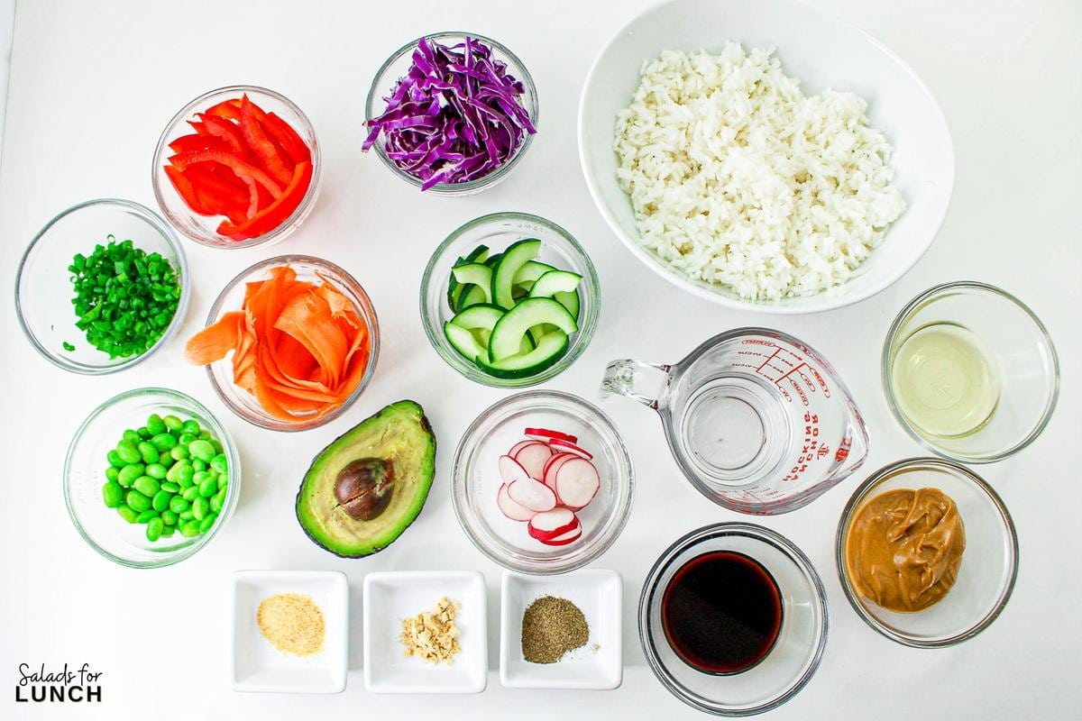 Overhead photo of ingredients used in a buddha bowl with peanut dressing, including: 2 cups cooked white rice,
½ cup shredded purple cabbage,
½ cup sliced red bell pepper,
½ cup sliced and halved cucumber, seeds removed,
⅓ cup shelled edamame,
⅓ cup sliced radishes,
⅓ cup sliced green onion,
1 avocado, sliced,
1 carrot, shaved,
1 teaspoon black sesame seeds, 
¼ cup creamy peanut butter,
¼ cup water,
2 tablespoons soy sauce,
2 tablespoons rice wine vinegar,
½ teaspoon ground ginger,
½ teaspoon garlic powder, and
½ teaspoon black pepper.