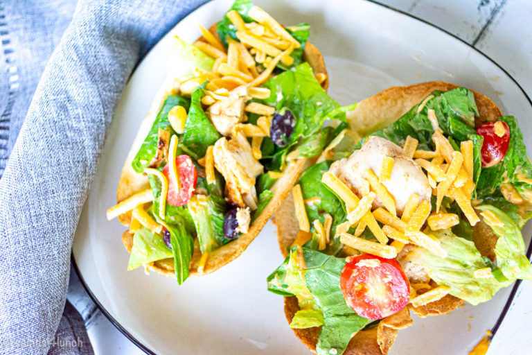 Two crispy edible taco bowls with chipotle chicken salad in them on a square white plate with black trim and a grey napkin beside the plate.