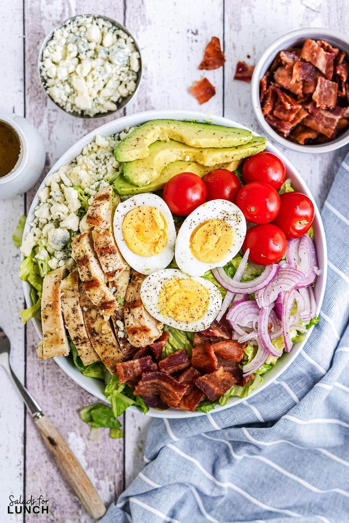 White bowl filled with cobb salad, layered with chicken, bacon, red onion, blue cheese, hard boiled eggs and cherry tomatoes on a blue and white striped napkin.