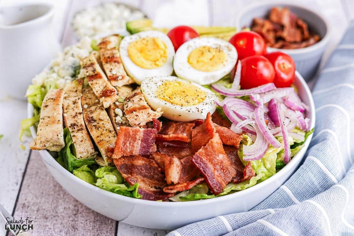 White bowl filled with cobb salad, layered with chicken, bacon, red onion, blue cheese, hard boiled eggs and cherry tomatoes on a blue and white striped napkin.