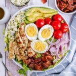 White bowl filled with cobb salad, layered with chicken, bacon, red onion, blue cheese, hard boiled eggs and cherry tomatoes on a blue and white striped napkin.