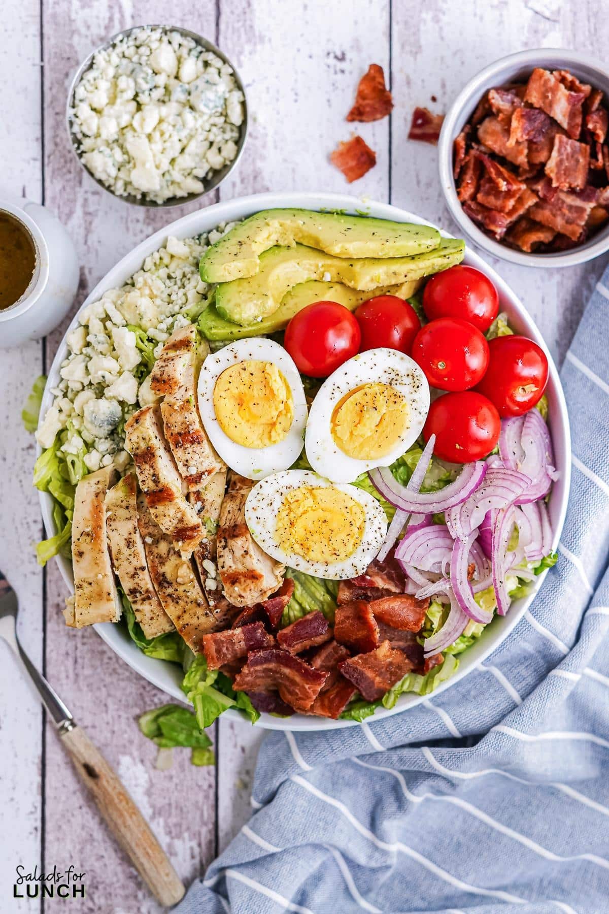 Classic Cobb Salad - Classic Cobb Salad 12 White bowl filled with cobb salad, layered with chicken, bacon, red onion, blue cheese, hard boiled eggs and cherry tomatoes on a blue and white striped napkin.