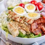Cobb salad layered with hard boiled eggs, cherry tomatoes, crispy bacon, chicken, lettuce and blue cheese in a white bowl.