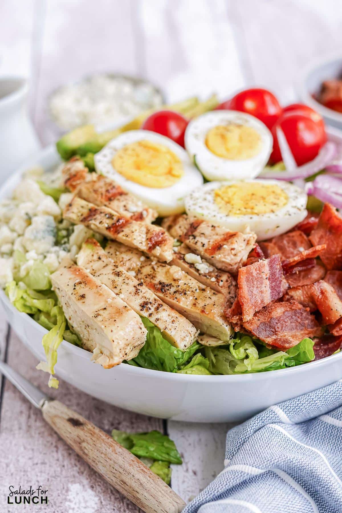 White bowl filled with cobb salad, layered with chicken, bacon, red onion, blue cheese, hard boiled eggs and cherry tomatoes on a blue and white striped napkin.