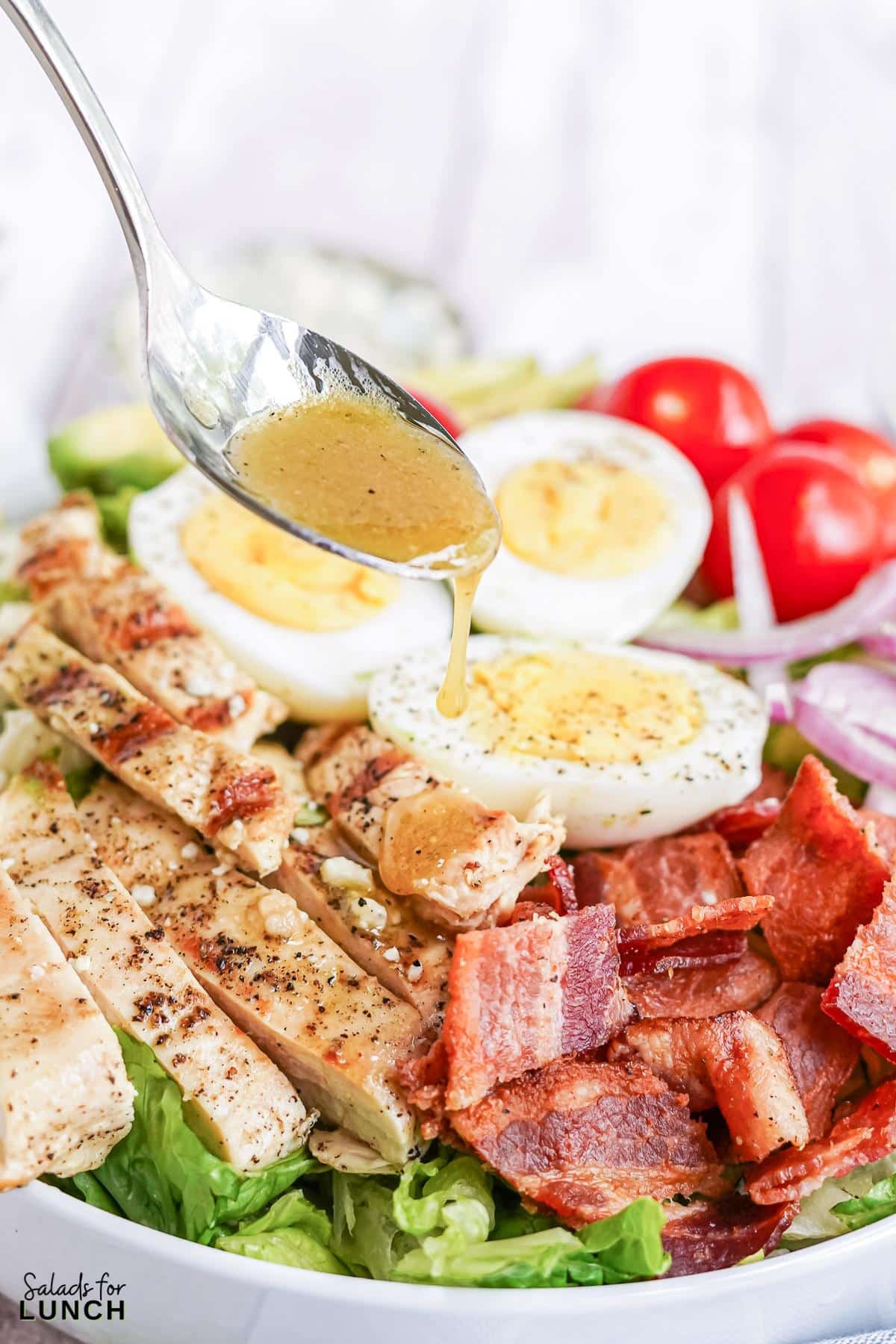 Salad dressing being drizzled onto a white bowl of cobb salad, layered with chicken, bacon, red onion, blue cheese, hard boiled eggs and cherry tomatoes on a blue and white striped napkin.
