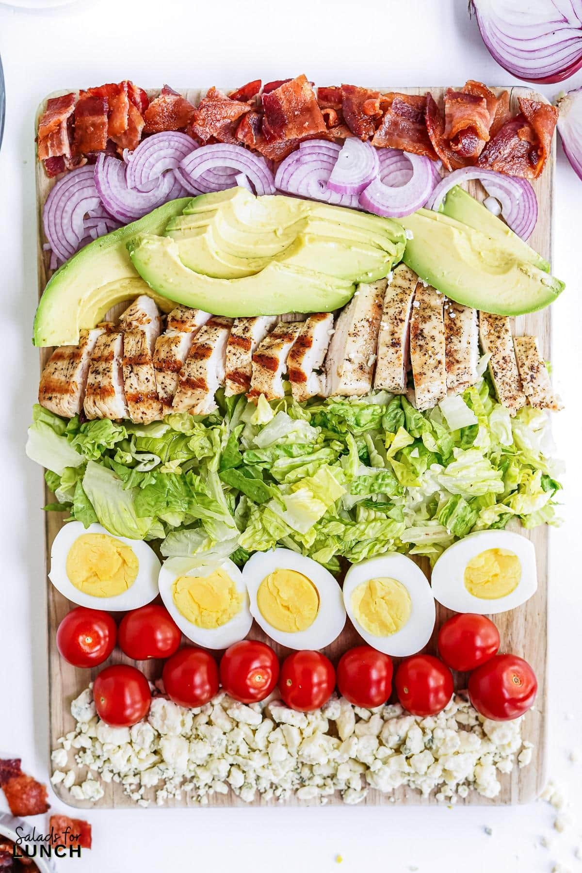 Cobb salad ingredients chopped and arranged on a wooden cutting board showing bacon, red onion, avocado slices, sliced chicken, cut romaine lettuce, hard boiled eggs, cherry tomatoes and blue cheese.