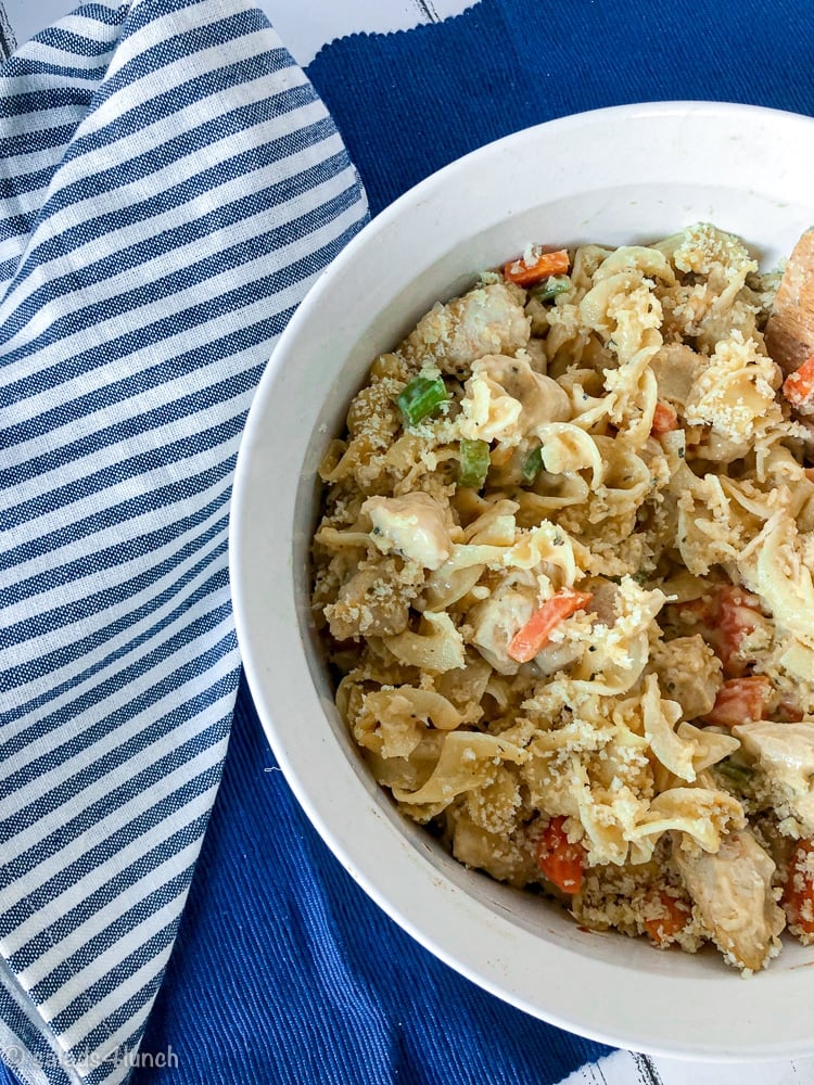 White casserole dish on a blue placemat with blue striped napkin with creamy chicken noodle casserole loaded with tender carrots and celery.