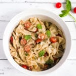 White bowl of bow tie feta bruschetta pasta salad with sprig of basil and roma tomatoes.