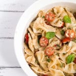 White bowl of bow tie feta bruschetta pasta salad with sprig of basil and roma tomatoes.
