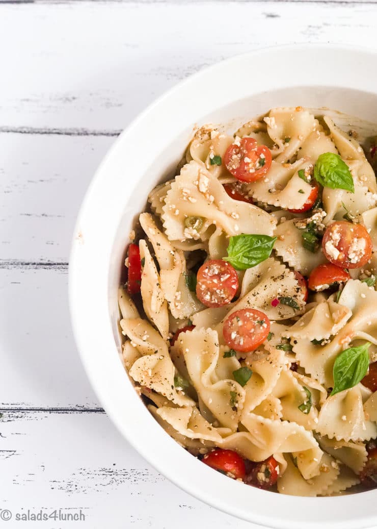 White bowl of bow tie feta bruschetta pasta salad with sprig of basil and roma tomatoes.