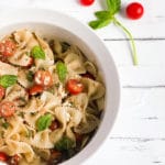White bowl of bow tie feta bruschetta pasta salad with sprig of basil and roma tomatoes.