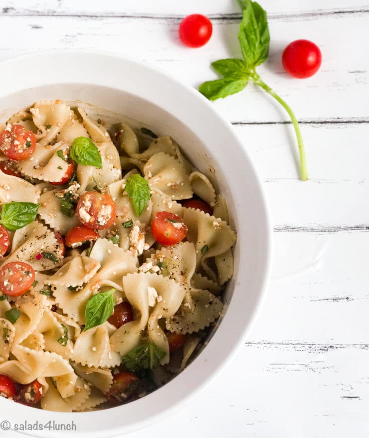 White bowl of bow tie feta bruschetta pasta salad with sprig of basil and roma tomatoes.