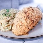 Close up of garlic parmesan baked chicken breast with a side of risotto on a white plate on a grey napkin.