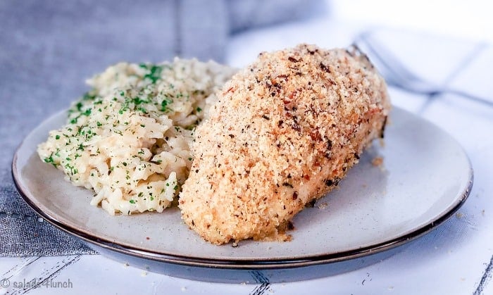 Close up of garlic parmesan baked chicken breast with a side of risotto on a white plate on a grey napkin.