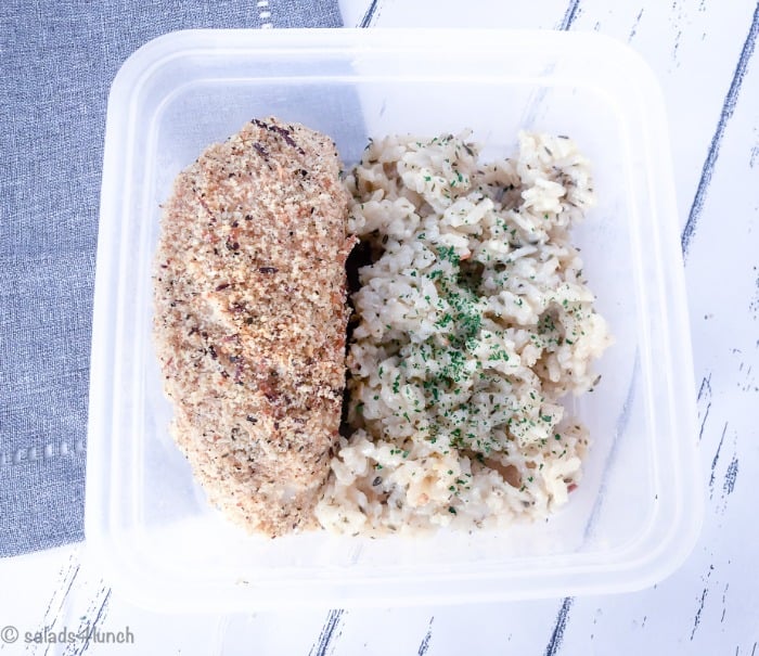 Close up of garlic parmesan baked chicken breast with a side of risotto in a meal storage container on a grey napkin.