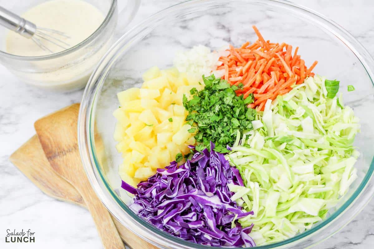 Clear glass mixing bowl with shredded green cabbage, red cabbage, carrots, cilantro and pineapple bits in it.