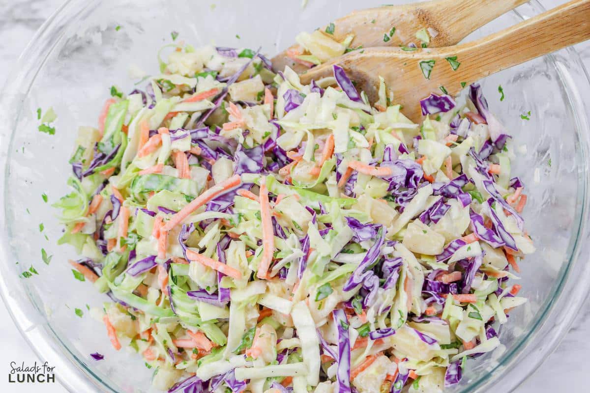 Coleslaw being mixed with dressing in a clear glass bowl.