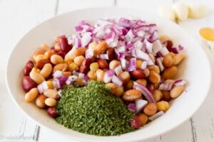 Close up of a white bowl on a white wood background with mixed beans, minced cilantro and red onion in it.