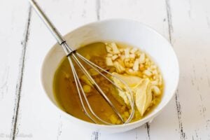 Close up of a white bowl with the dressing ingredients for mixed bean salad: Garlic, olive oil, dijon mustard, honey and white wine vinegar with a whisk in the bowl.