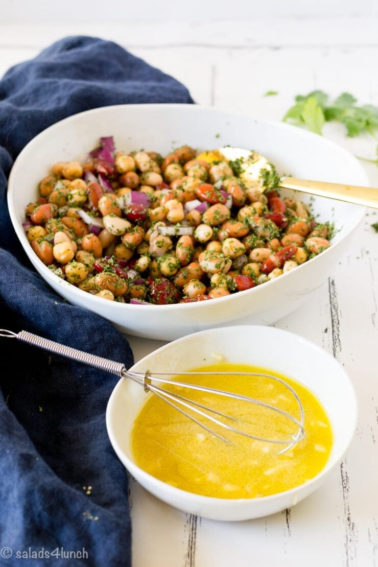 Two white bowls on a blue napkin, one bowl has mixed bean salad in it, and the other has the dressing with a whisk in it ready to mix together.