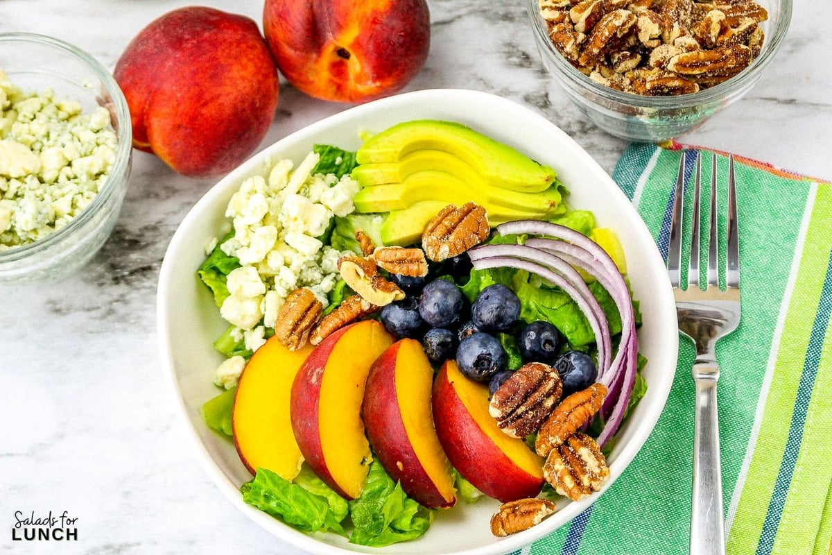 Summer peach salad with fresh peaches, blueberries, onion, romaine lettuce, pecans and blue cheese in a square white bowl with fresh mint dressing in the background on a green napkin and a white marble surface