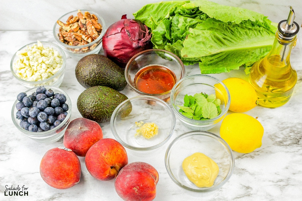 Ingredients for summer peach salad on a white marble surface: Blue cheese, roasted pecans, red onion, avocado, romaine lettuce, olive oil, lemons, mint, garlic, peaches, and blueberries.