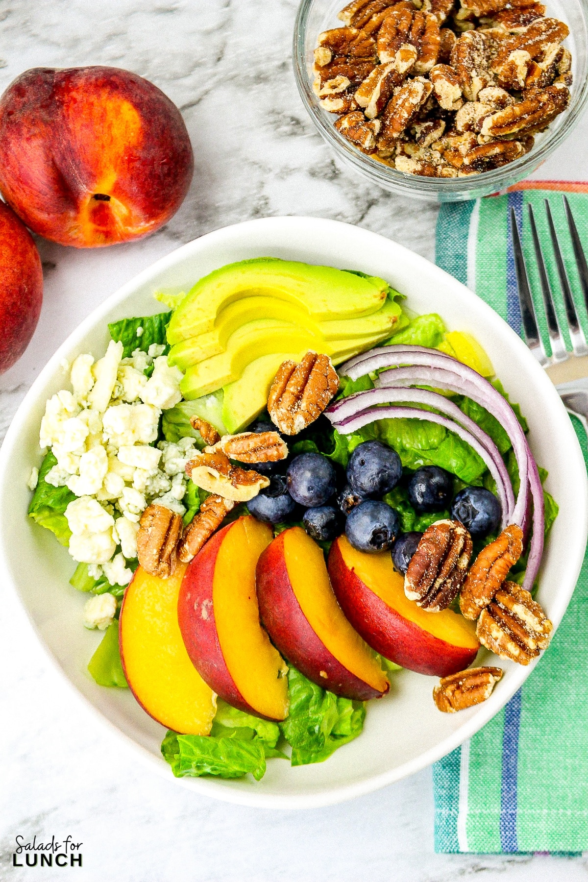 Summer peach salad with fresh peaches, blueberries, onion, romaine lettuce, pecans and blue cheese in a square white bowl with fresh mint dressing in the background on a green napkin and a white marble surface